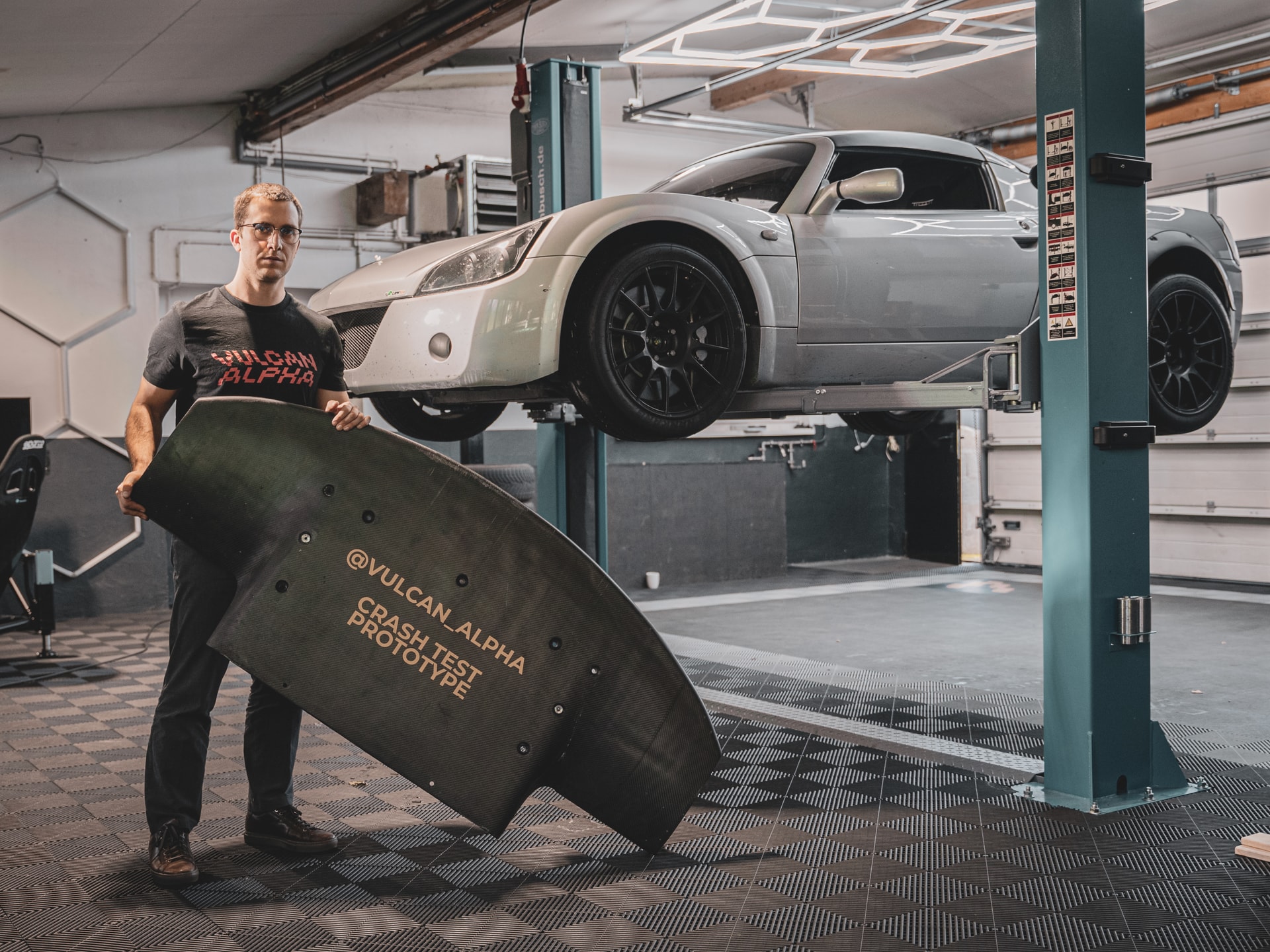 Man holding a crash test prototype of carbon fiber aerodynamic part for racing cars with an elevated fancy silver sport car in the background