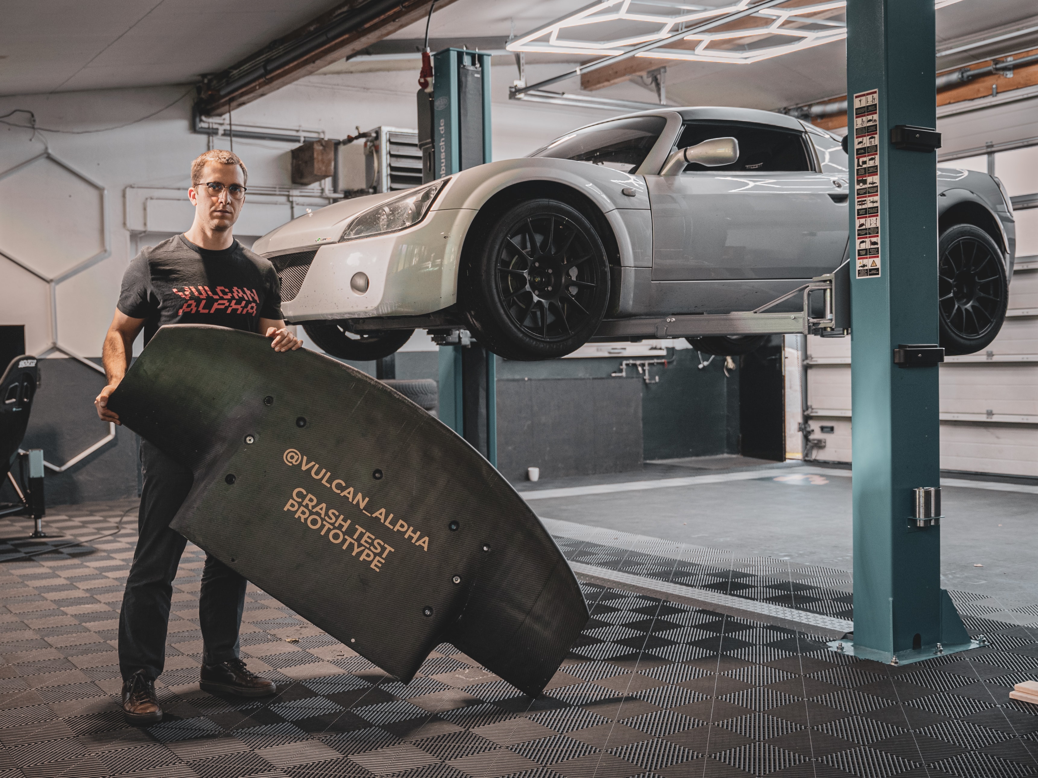 Man holding a crash test prototype of carbon fiber aerodynamic part for racing cars with an elevated fancy silver sport car in the background