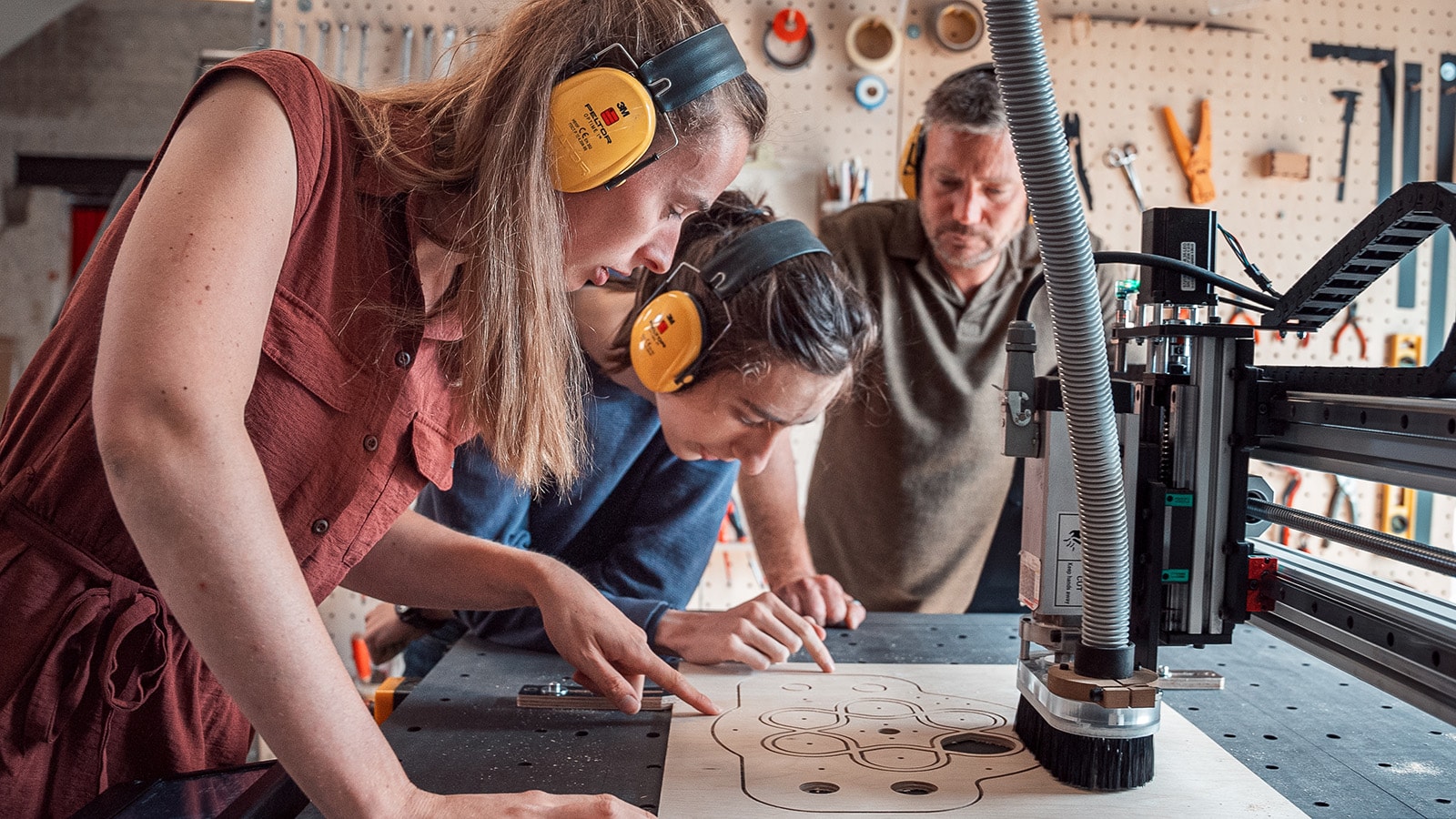 Fablab members wearing noise protection and observing a wood part cut on a CNC milling machine